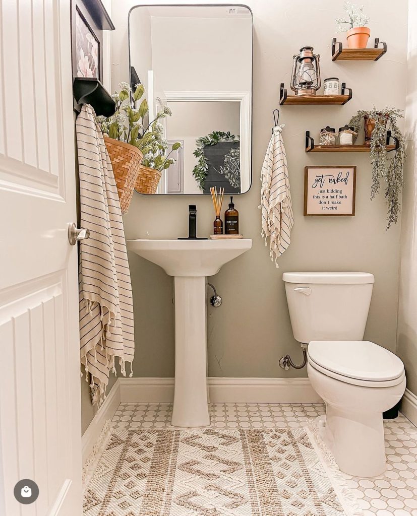 Neutral boho half bathroom with small floating wood shelves styled with a vintage lantern, terracotta pot, trailing faux plant, and a small wooden sign, plus a tasseled Turkish towel on the door and a woven rug.