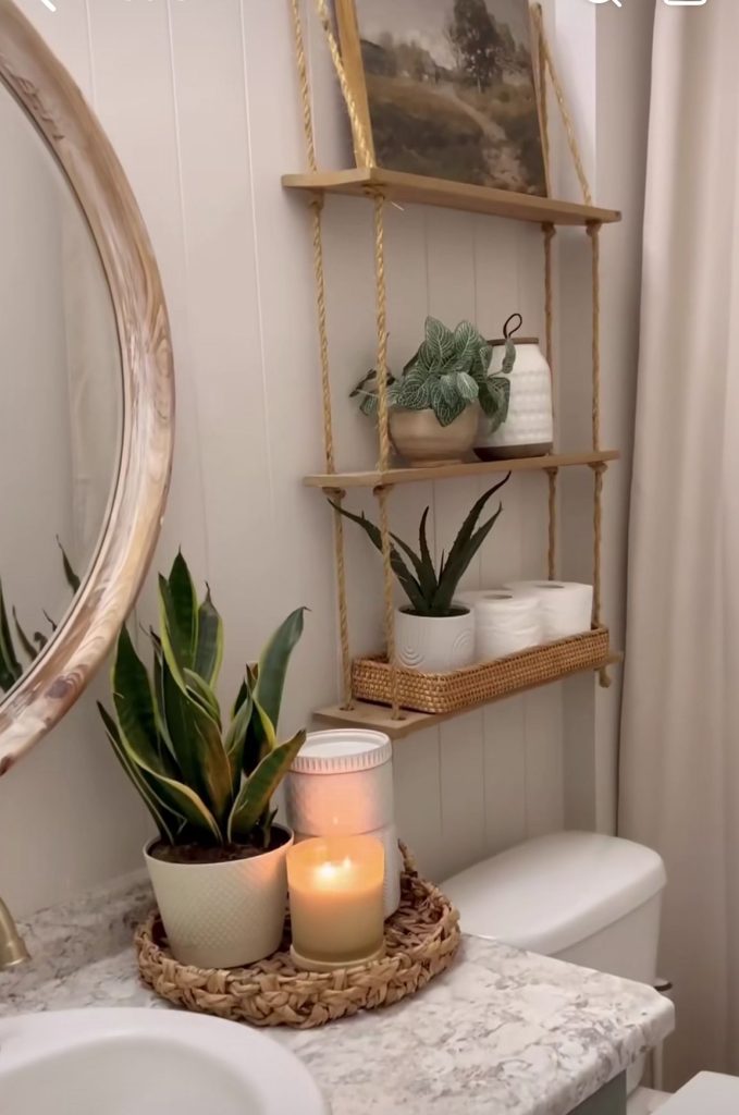 Neutral bathroom with a three tier rope hanging shelf holding a fern, snake plant, white vase, and a woven rattan tray with toilet rolls, plus a seagrass tray on the tank with a plant and candle.