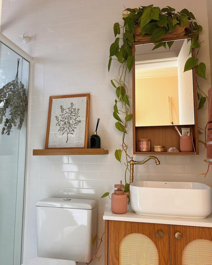 White subway tile boho bathroom with a wooden framed botanical art print on a floating shelf, a trailing pothos hanging from a rattan mirror cabinet, dried eucalyptus from the shower screen, and a cane fronted vanity unit.