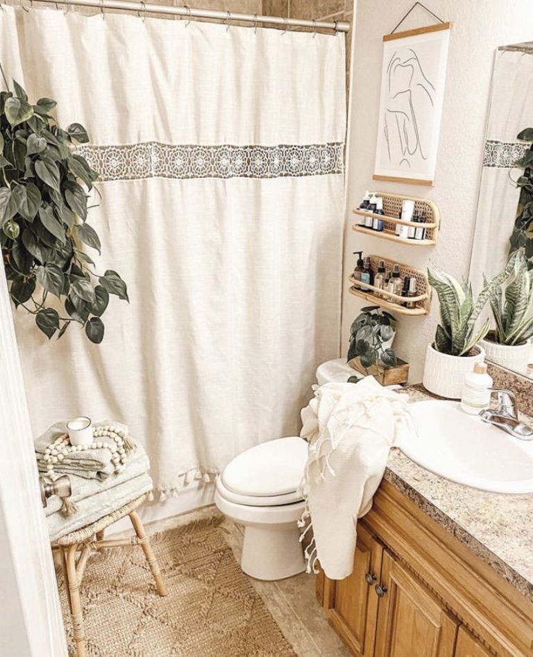 Warm neutral boho bathroom with a cream linen look shower curtain with a crochet detail border and tassels, a rattan stool with a wooden bead garland, rattan wall shelves with beauty products, and a line art poster with a wood hanger.