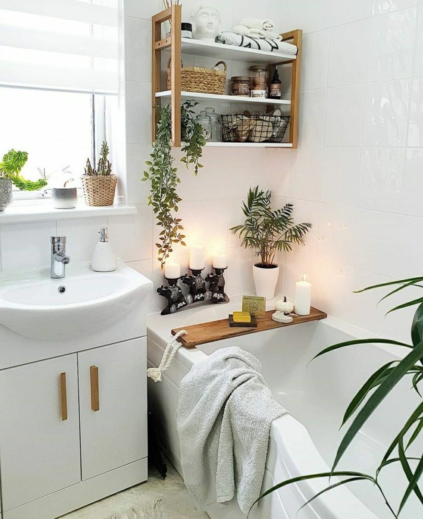 White bathroom with a rustic wooden bath tray holding candles and soap across the tub, a row of pillar candles on dark holders on the tub edge, a bamboo framed wall shelf above with baskets and a buddha figurine, and plants on the windowsill.