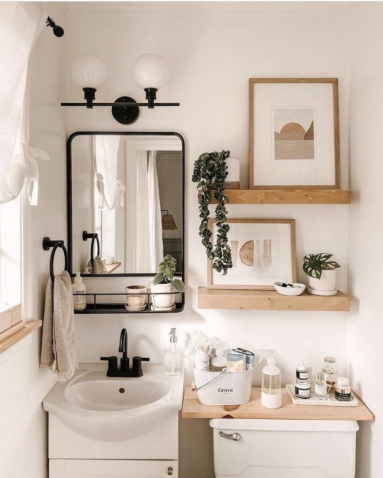 Bright white bathroom with two floating wood shelves styled with framed boho abstract art prints, a trailing plant, and a small monstera in a white pot, plus a black framed mirror with a shelf below and a dual globe wall sconce light.