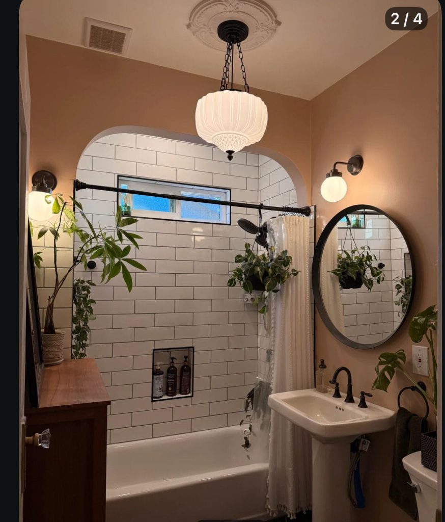 Bathroom with multiple leafy plants on surfaces and hanging near the shower, a vintage globe pendant light, and a round black framed mirror above a pedestal sink.
