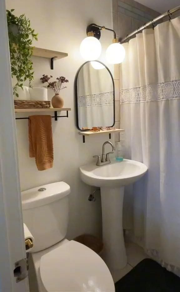 Small neutral bathroom with an arched black frame mirror above the sink, a double sconce light, a wooden floating shelf with a terracotta vase and dried flowers, and a lace trim shower curtain.