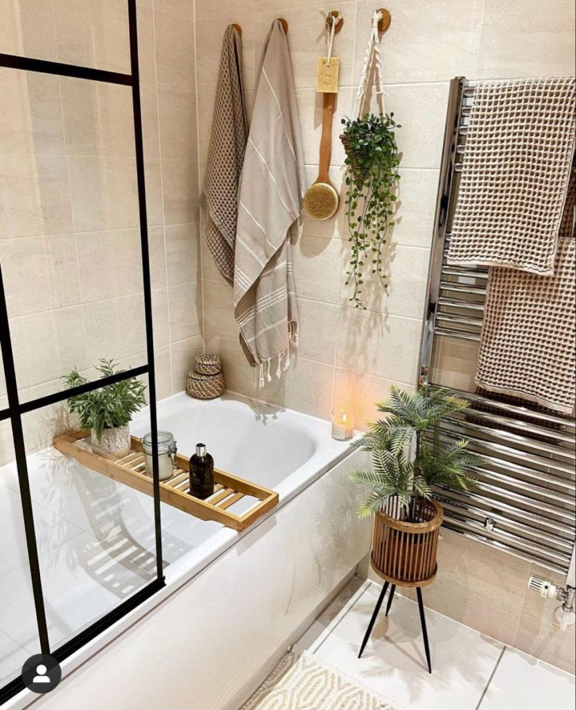 Neutral boho bathroom with a bamboo bath tray on the tub holding a plant and products, textured towels hanging from wooden wall hooks, a string of pearls plant in a macrame hanger, and a rattan plant stand.