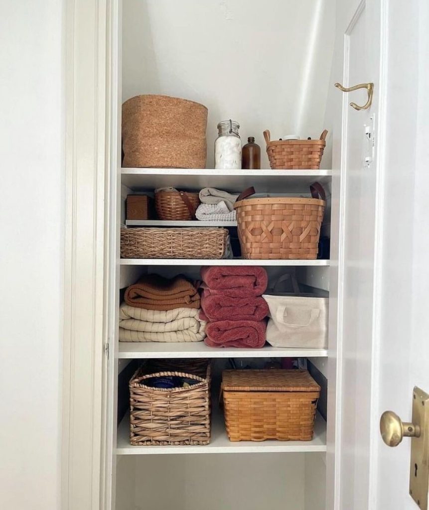 Open white linen closet with multiple woven wicker and leather handled baskets on shelves, rolled towels in warm tones, and a cork storage pot on the top shelf.