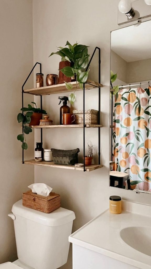 Small bathroom with a three tier metal and wood ladder style wall shelf above the toilet, styled with plants in terracotta pots, amber glass bottles, a woven basket, and a fruit print shower curtain to the side.