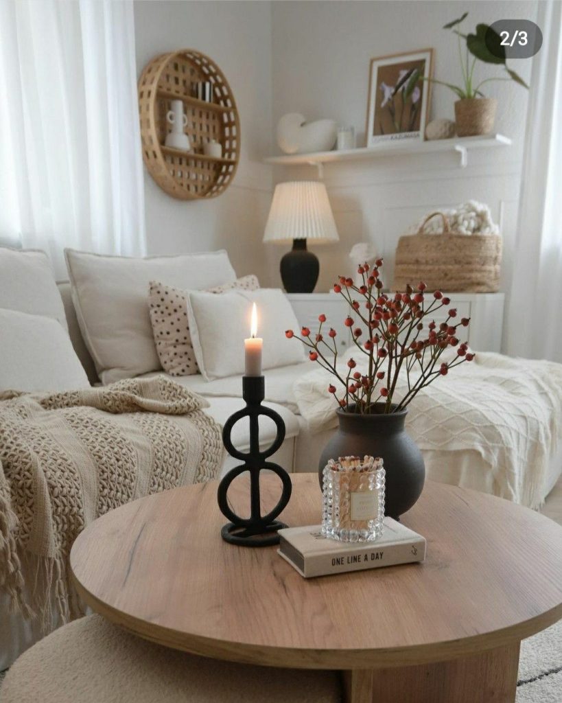 Round wood coffee table styled with a black sculptural candle holder with a lit taper candle, a black vase with red berry stems, and a glass candle on a book.