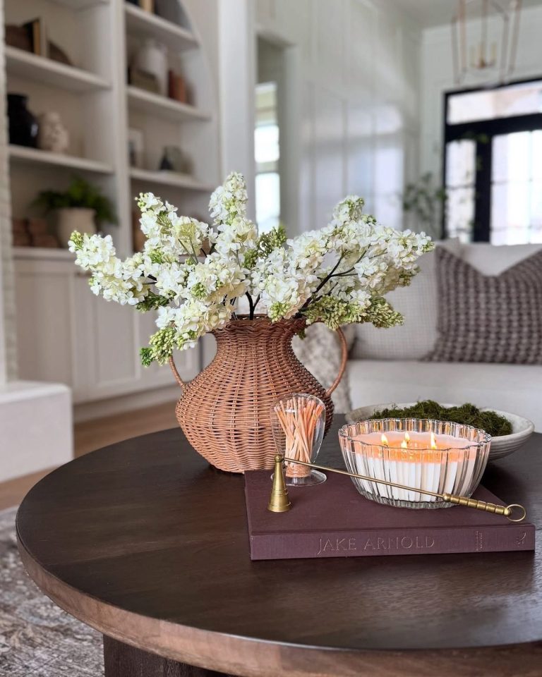 Dark wood round coffee table with a woven rattan vase holding white faux lilac stems, a multi-wick candle in a glass ribbed bowl, a glass matchstick holder, and a decorative book underneath.