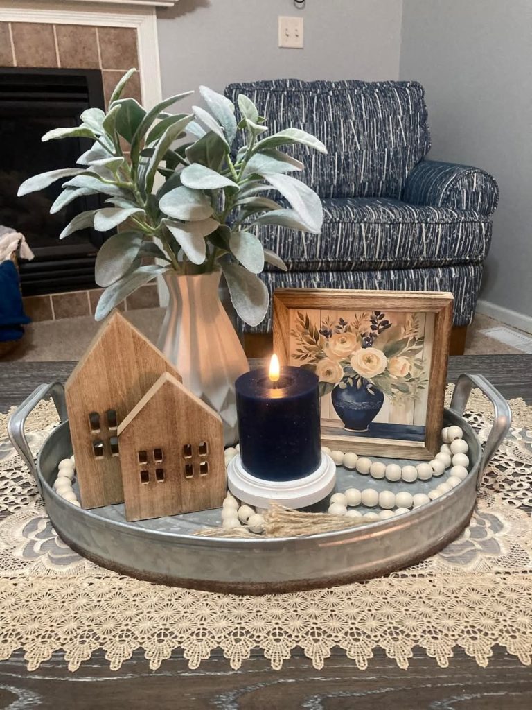 Oval galvanized metal tray on a dark wood coffee table with a crochet lace runner, styled with wooden house figurines, a terracotta vase with lamb's ear stems, a dark pillar candle, a small framed floral print, and a wood bead garland.