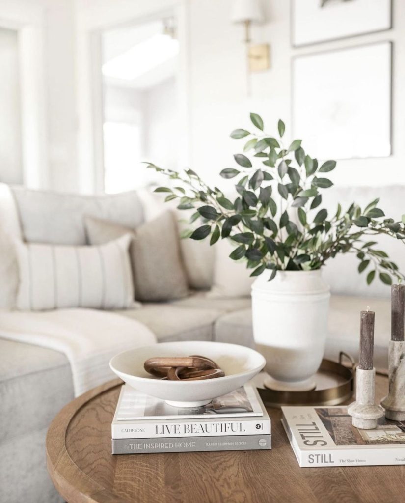 Round oak coffee table styled with a large white ceramic vase holding tall green branches, two stacks of white coffee table books topped with a white bowl holding wooden objects, and slim stone candle holders.