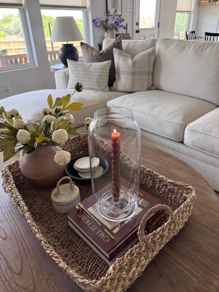 Rectangular seagrass tray on a wood coffee table with a glass hurricane holding a lit burgundy taper candle on books, a round pink vase with white faux ranunculus, a small ceramic lidded box, and a dark plate with marble pebbles.