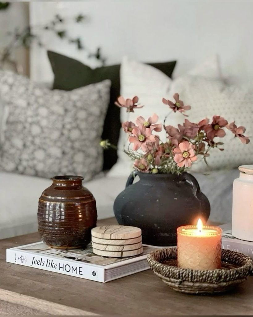 Wood coffee table styled with a dark matte handled ceramic jug holding pink cosmos stems, a small ribbed brown pottery vase, a stack of books, marble coasters, and a glowing textured glass candle in a small wicker dish.