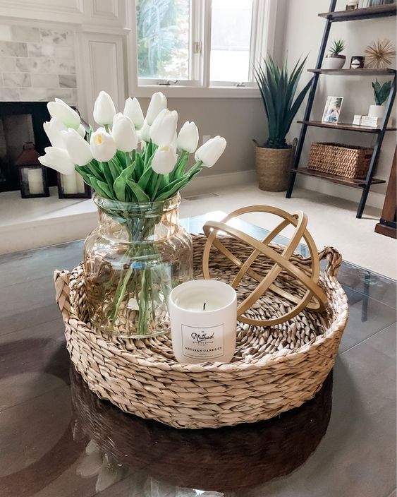 Round wicker tray on a glass coffee table with a clear glass vase holding white faux tulips, a small white jar candle, and a natural rattan orb sculpture.