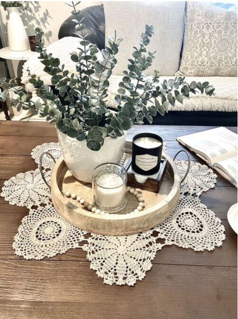 Crochet lace doily on a dark wood coffee table with a round wood tray holding a white ceramic pot with faux eucalyptus, a glass votive candle, a black jar candle on a small wood crate, and a wood bead garland.