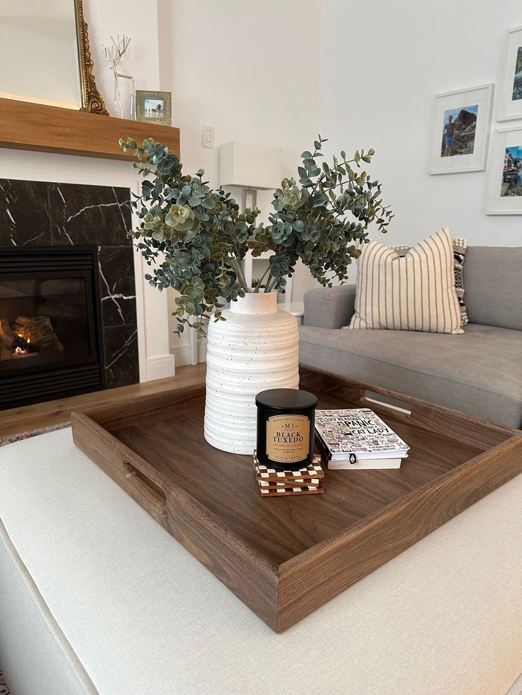 Wood serving tray on a cream ottoman with a white ribbed vase holding faux eucalyptus branches, a black candle, and a checkered coaster set.