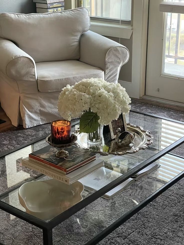 Glass and black metal coffee table styled with a vintage silver ornate tray holding a clear glass vase with white hydrangeas, a small silver swan figurine, and an amber glass candle in a footed holder, with stacked coffee table books and a white decorative dish in the foreground.