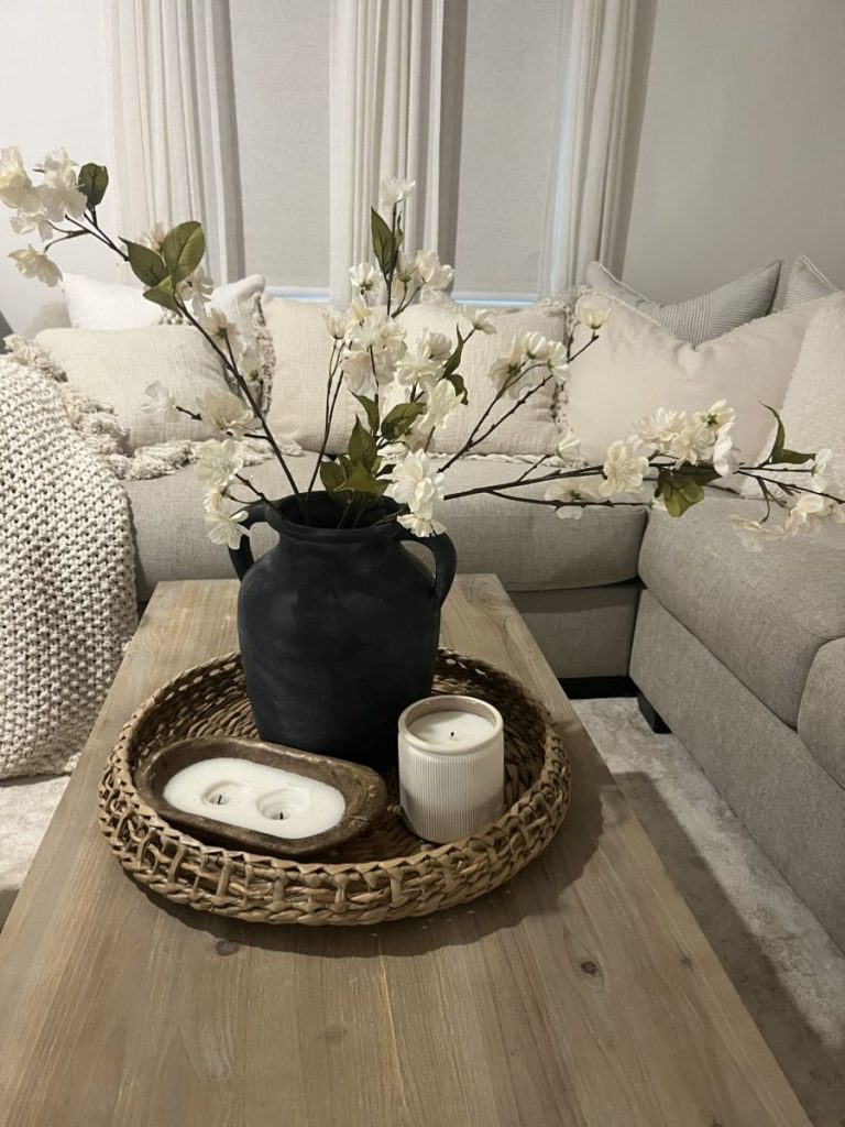 Round wicker tray on a wood coffee table with a dark ceramic jug holding white blossom stems, a wooden dough bowl with tealights, and a small white ribbed candle.
