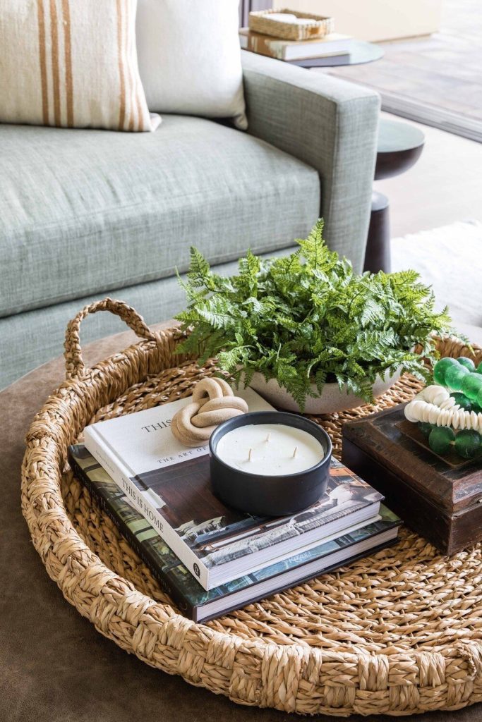 Large round seagrass tray on a coffee table with stacked home decor books, a black bowl candle on top, a small potted fern in a white bowl, and a knotted ceramic object.
