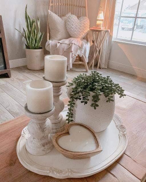 Distressed white decorative tray on a wood coffee table styled with two white pillar candles on turned wood candlesticks, a succulent in a white ceramic pot, and a wooden heart shape.