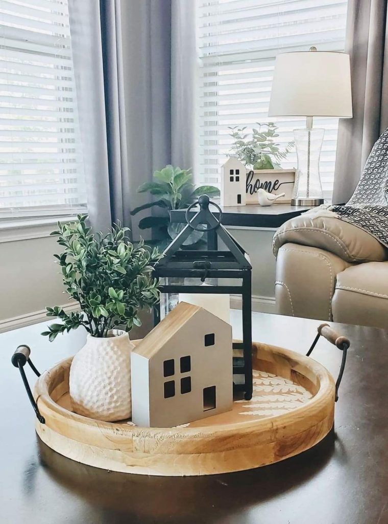 Round mango wood tray on a dark coffee table styled with a black metal lantern, a white house figurine, and a small textured white vase with faux boxwood greenery.