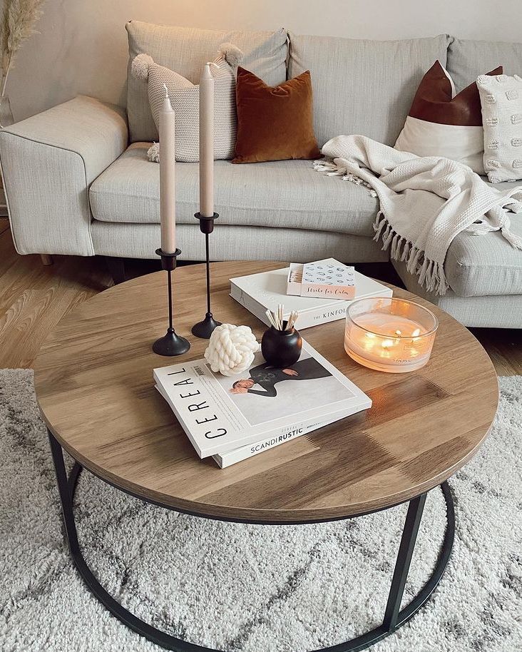 Round wood and black metal coffee table styled with two pink taper candles in black holders, stacked white books, a small knotted white sculpture, a black matchstick holder, and a glass candle bowl with fairy lights.