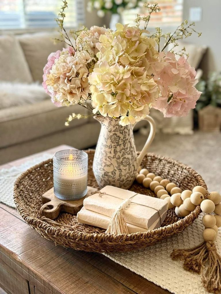 Wicker tray on a wood coffee table with a floral printed ceramic pitcher holding blush and cream faux hydrangeas, a glass votive candle, kraft-wrapped books, and a wooden bead garland.