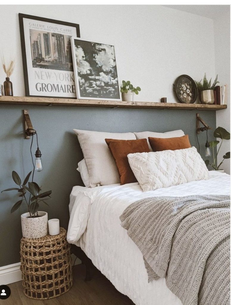 Rustic wooden shelf above bed with leaning art prints, small plants, and Edison bulb pendant lights against a sage green accent wall.