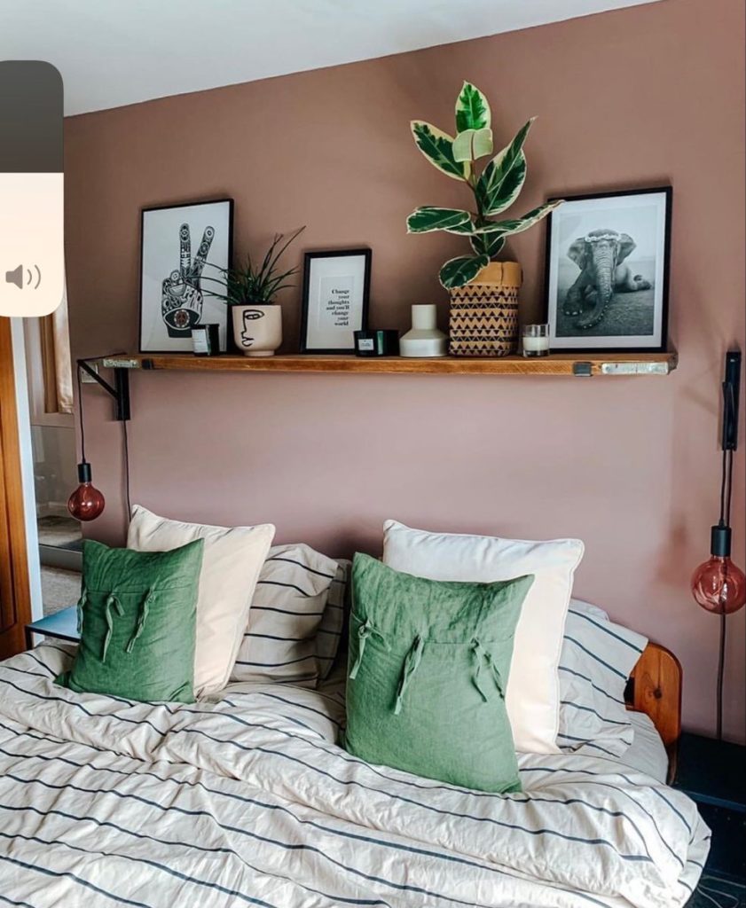 Rustic wooden shelf on a dusty rose pink wall above a bed with striped bedding and green linen cushions, styled with black framed art prints, a large variegated rubber plant, face planter, candles, and small ceramic decorative objects, with hanging red globe pendant lights on either side.