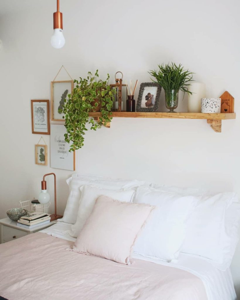 Rustic wooden shelf above a bed styled with large trailing variegated pothos plant, a copper lantern, reed diffuser, framed photo, green plant in a glass vase, and a small decorative house, with additional wall art hanging below on either side.