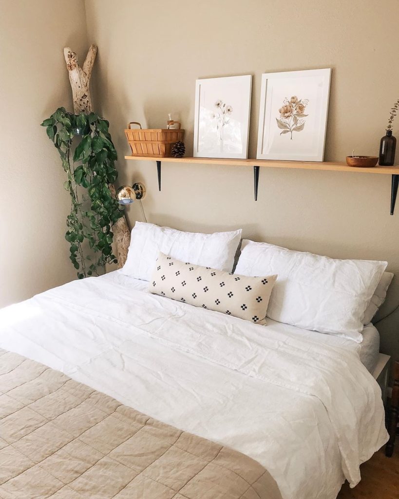 Natural wood shelf on a warm greige wall above a bed with crisp white linen, styled with two soft botanical watercolor prints in white frames, a small woven basket, a pinecone, and a wooden bowl, with a large trailing pothos climbing a piece of driftwood in the corner.