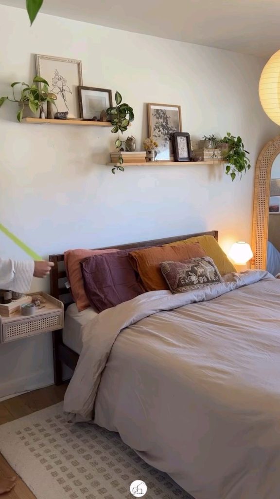 Natural wood shelf above a dark wooden bed frame styled with a mix of small framed art prints, trailing and potted plants including a pothos and small succulents, and a cane arch mirror leaning against the wall to the side.