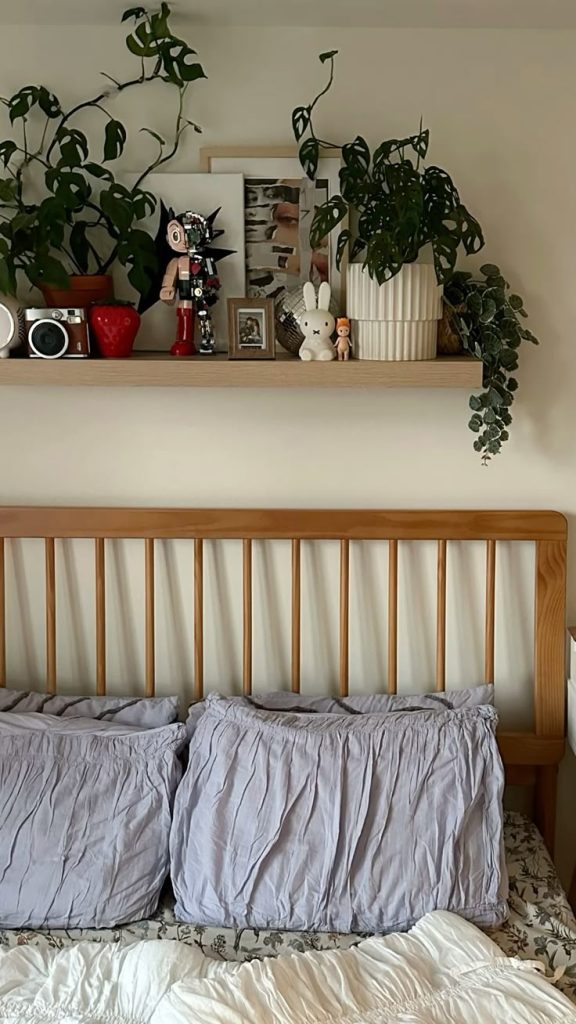 Light wood shelf above a spindle wooden bed headboard overflowing with large monstera and pothos trailing plants, photo frames, a film camera, a white bunny figurine, and small collectible ornaments.