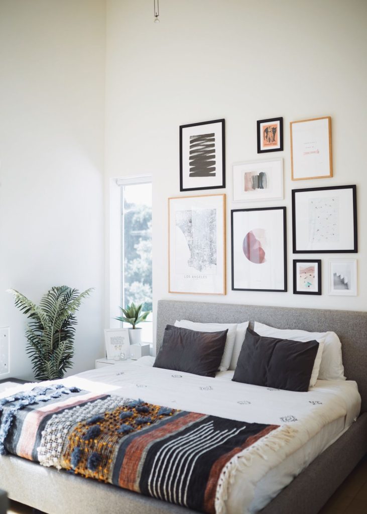 Airy modern bedroom with high ceilings and a mixed gallery wall of seven framed prints in black and natural wood frames above the bed, featuring abstract watercolor art, a city map print, and botanical illustrations.