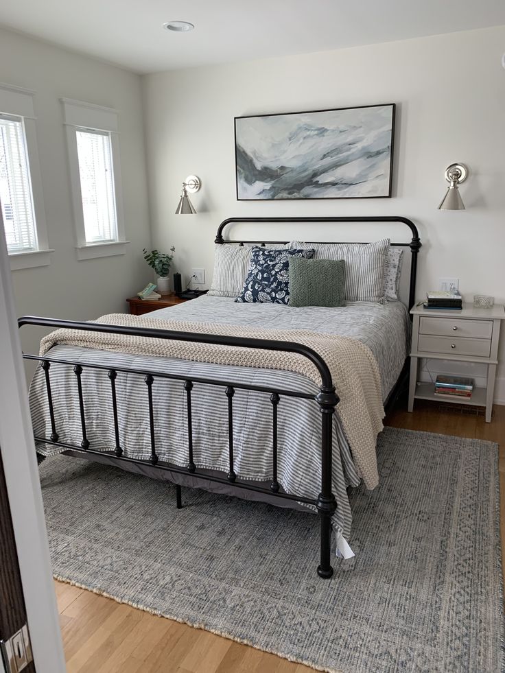Small neutral bedroom with a single large horizontal abstract blue and grey canvas painting hung above a black iron bed, flanked by two chrome wall sconces.