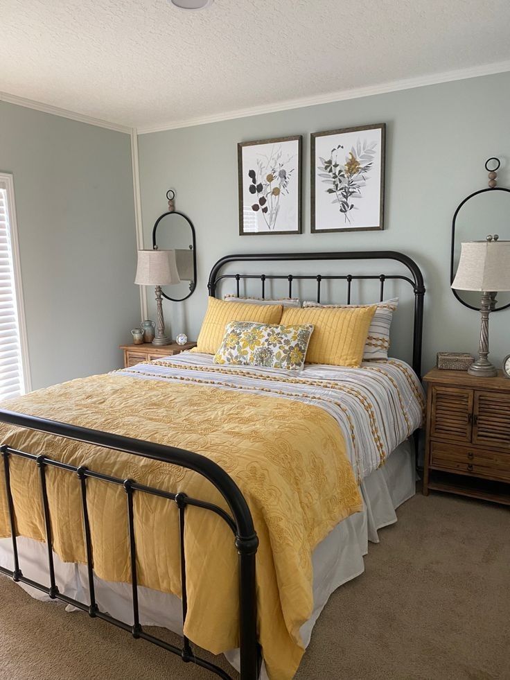 Light sage green bedroom with a black iron bed and two matching framed botanical prints side by side above the headboard, with warm wood nightstands and yellow bedding accents.