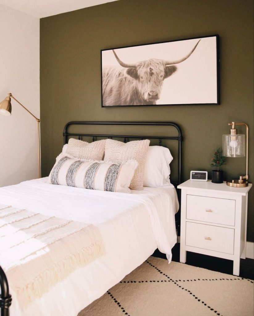 Cozy bedroom with a deep olive green accent wall behind a black iron bed, a single large framed horizontal canvas of a Highland cow print above the headboard, and a white nightstand with gold hardware.