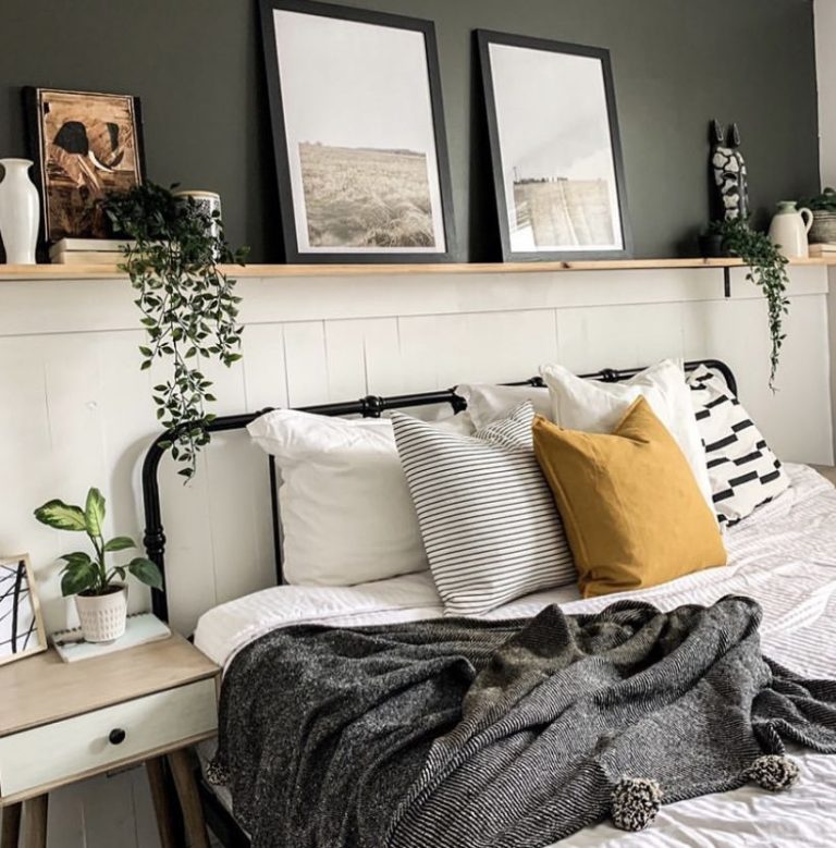 Moody bedroom with a dark olive green wall and white shiplap lower half, a long natural wood floating shelf styled with framed landscape prints, trailing pothos plants, and small ceramic decor objects above a black iron bed.