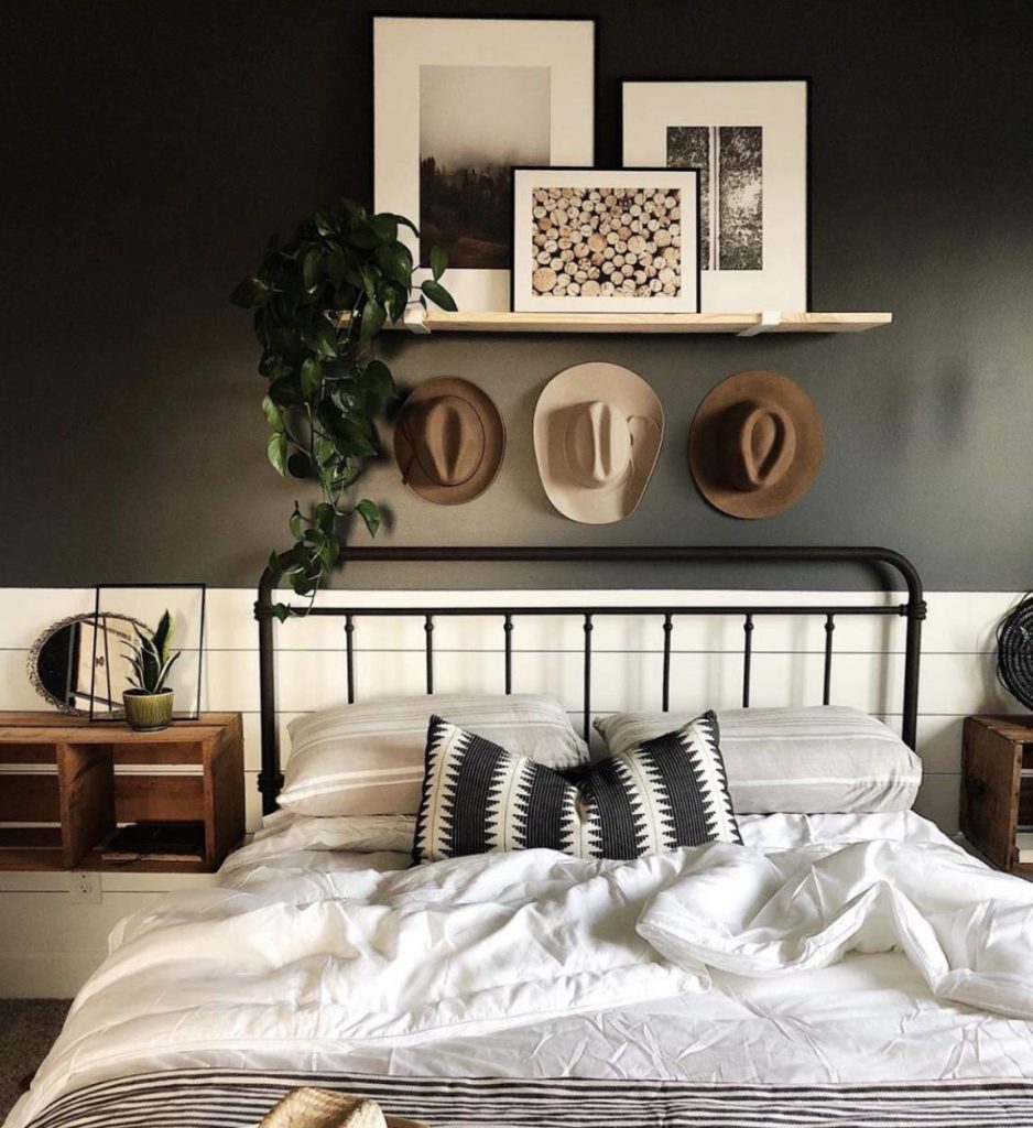 Dark charcoal bedroom wall with a floating natural wood ledge shelf above a black iron bed, styled with layered landscape prints and a trailing plant, with three felt wide-brim hats displayed on hooks below the shelf.