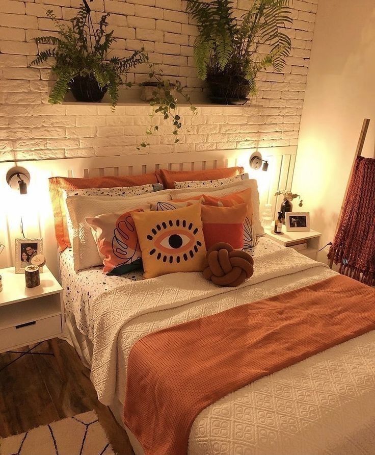 Warm cozy bedroom with a whitewashed brick wall behind the bed, a floating shelf with dark potted ferns and plants trailing over the edges, warm Edison bulb wall sconces on either side, and rust orange bedding.