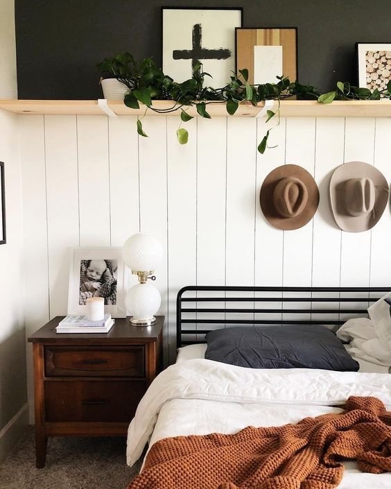 Minimalist modern bedroom with a white shiplap feature wall, a slim natural wood floating shelf above the bed styled with a cross print, a wooden frame, and a trailing plant, and two felt hats hung on hooks on the wall below.