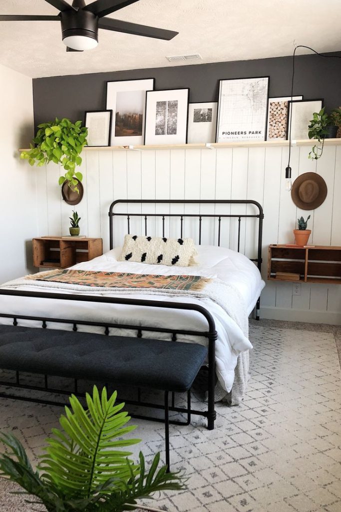 Modern farmhouse bedroom with a long wooden ledge shelf above a dark accent wall, styled with framed black and white prints, greenery, and hats hanging on shiplap below.