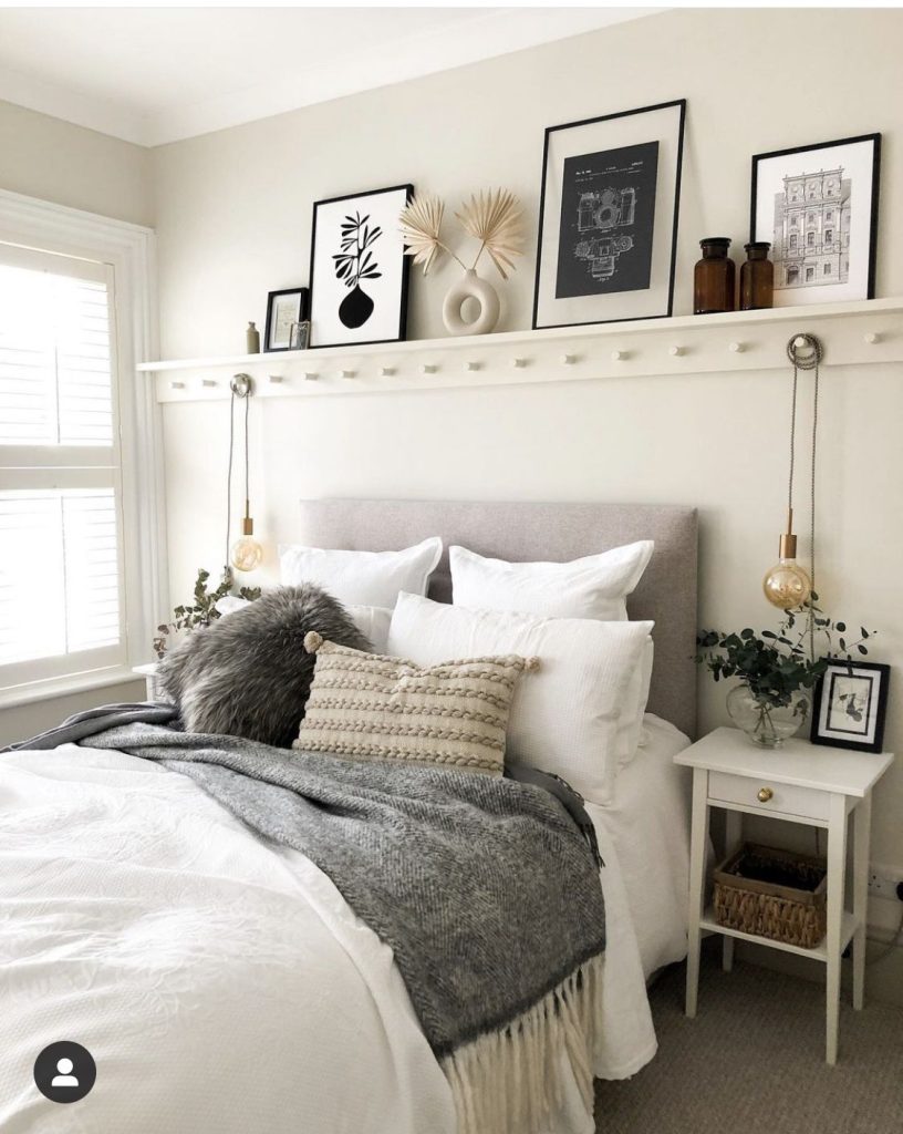 Soft neutral bedroom with a white picture ledge shelf above the bed styled with layered black framed prints, dried palm fans, a ceramic donut vase, and amber glass bottles.