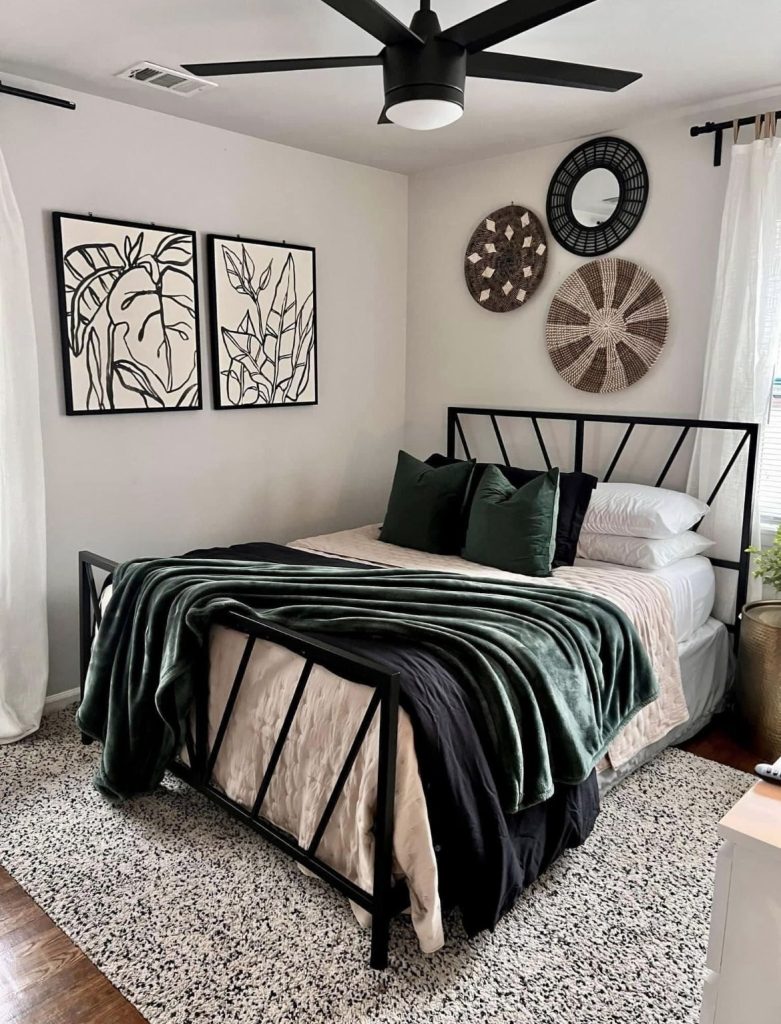 Neutral modern bedroom with a black metal bed frame, two large framed botanical line art prints on one wall, and a cluster of three woven rattan baskets and a round mirror on the adjacent above bed wall.