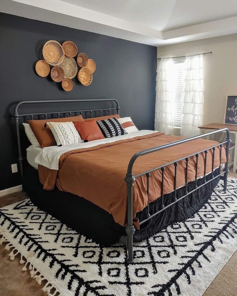 Moody boho bedroom with a dark navy accent wall above the bed styled with a cluster of assorted woven rattan and wicker baskets in varying sizes, with a rust orange bedspread and geometric patterned rug.