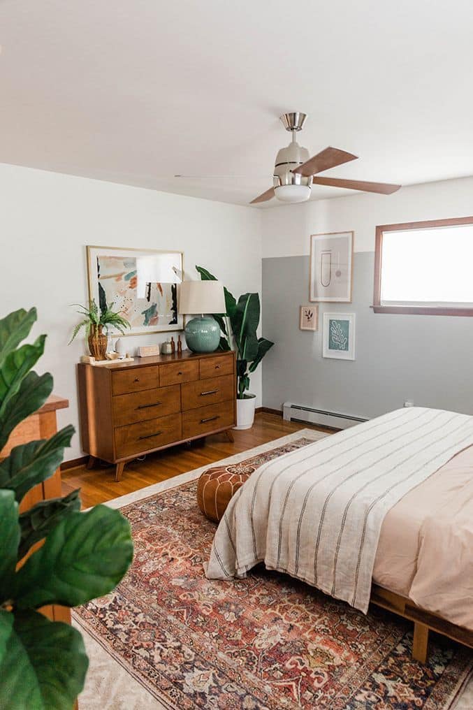 Boho bedroom with a two-tone grey and white accent wall, warm wood dresser, vintage rug, and large houseplants
