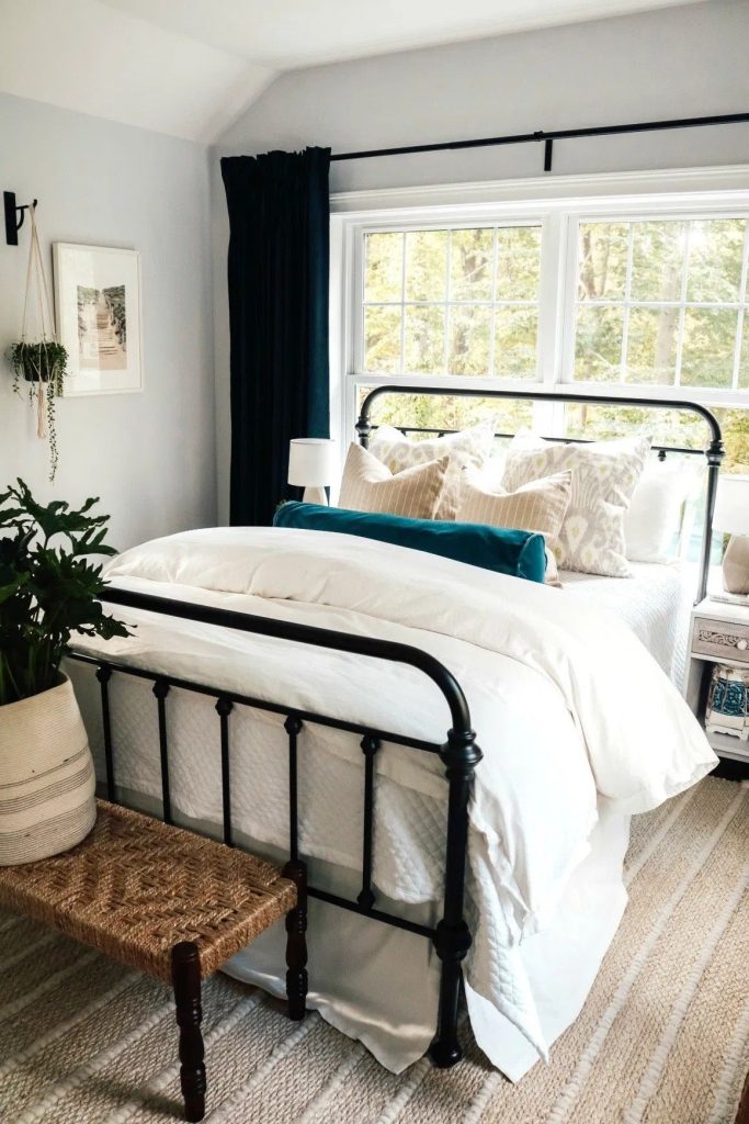 Boho bedroom with a black iron bed frame, white bedding, teal velvet bolster pillow, and a rattan bench