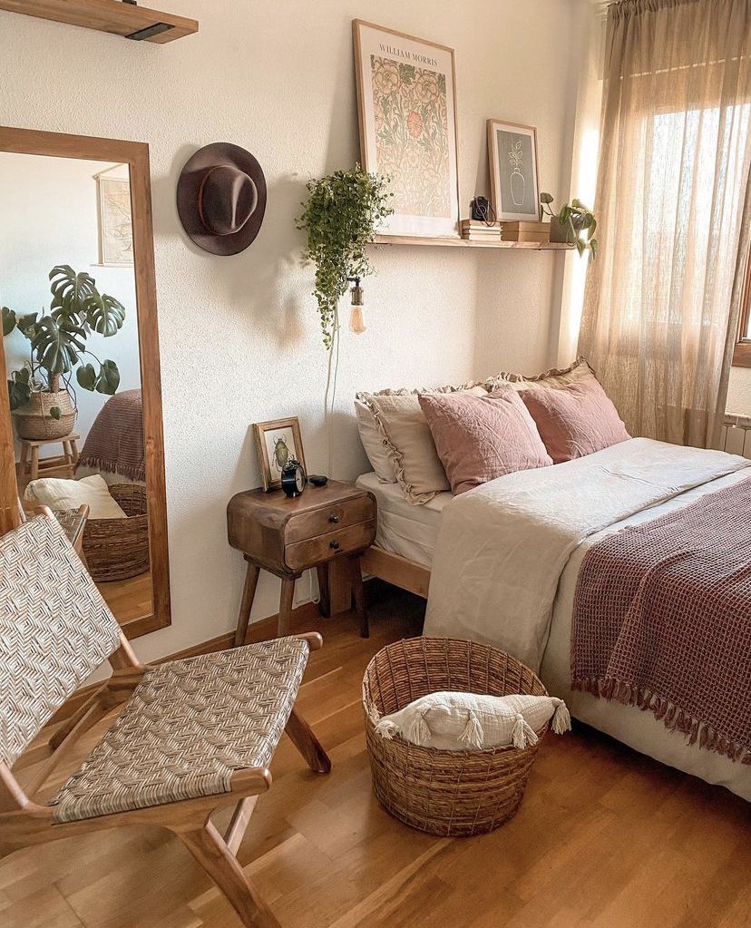 Small boho bedroom with warm pink bedding, a woven rattan chair, wooden nightstand, large floor mirror, and trailing plants