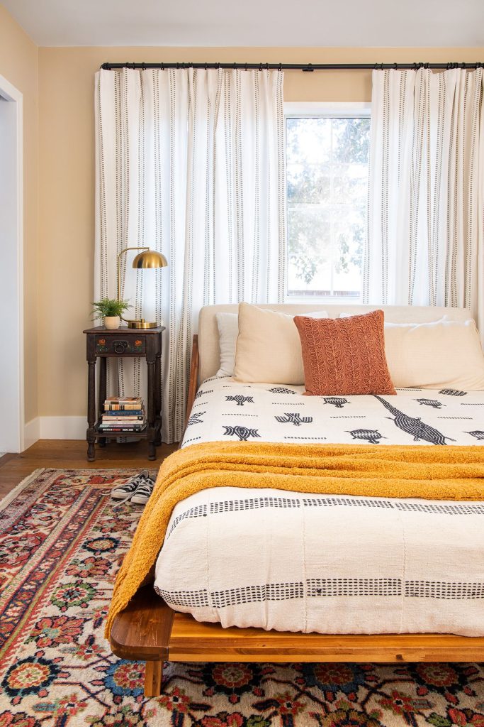 Boho bedroom with wide striped curtains hung behind the bed, a vintage Persian rug, golden throw, and dark wooden nightstand