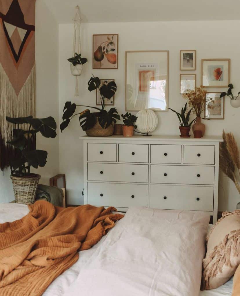 Boho bedroom with a white dresser styled with monstera plants, dried grasses, gallery wall art, and woven baskets